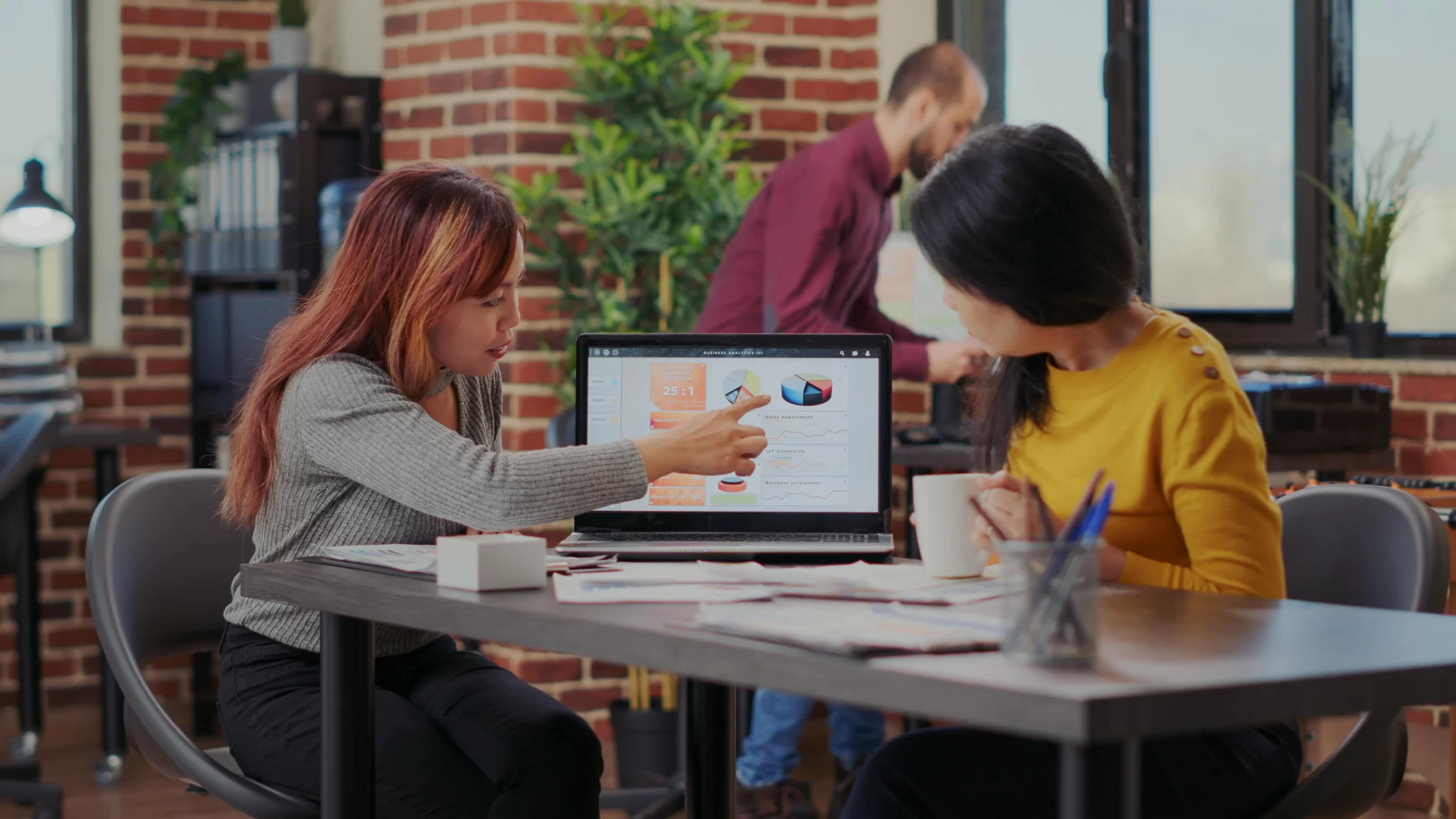 Two women in an office discussing Google Ads report through charts and graphics on a laptop screen.