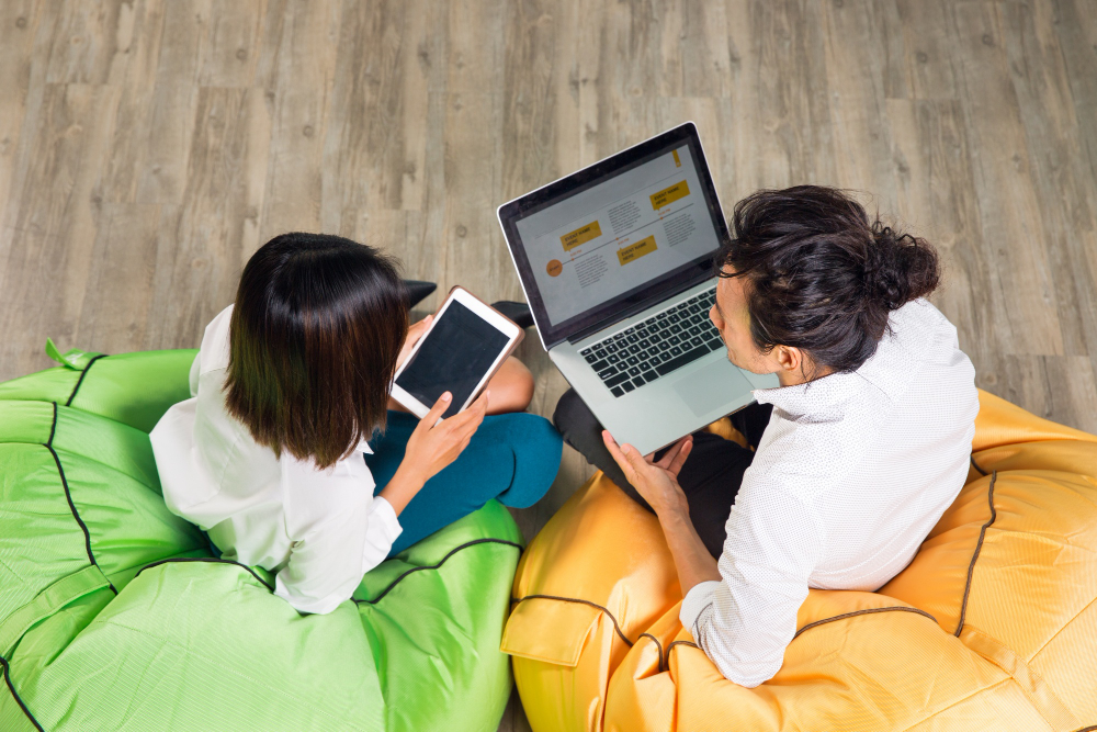 A high-angle shot of a man and a woman sitting on a beanbag chair holding smart gadgets while having an interactive conversation.