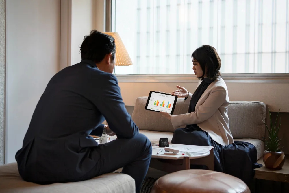 Is Your Business Ready to Grow Using a Google Ads Agency? 1 A businesswoman sitting on a sofa holding a tablet that has a colourful bar chart on screen, presenting data to a businessman in front of her.