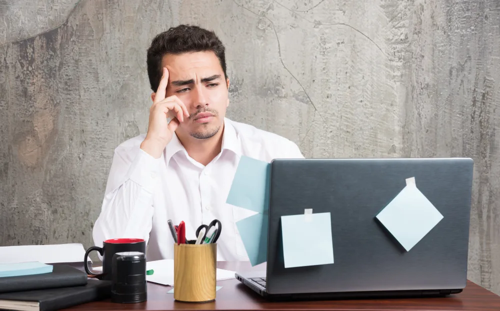 A concerned man in a white button-down shirt sits at a desk, resting his head on his hand while staring at the laptop screen covered in sticky notes.